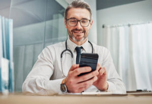 Healthcare, smile and doctor with smartphone at desk for wellness research, medical app and online