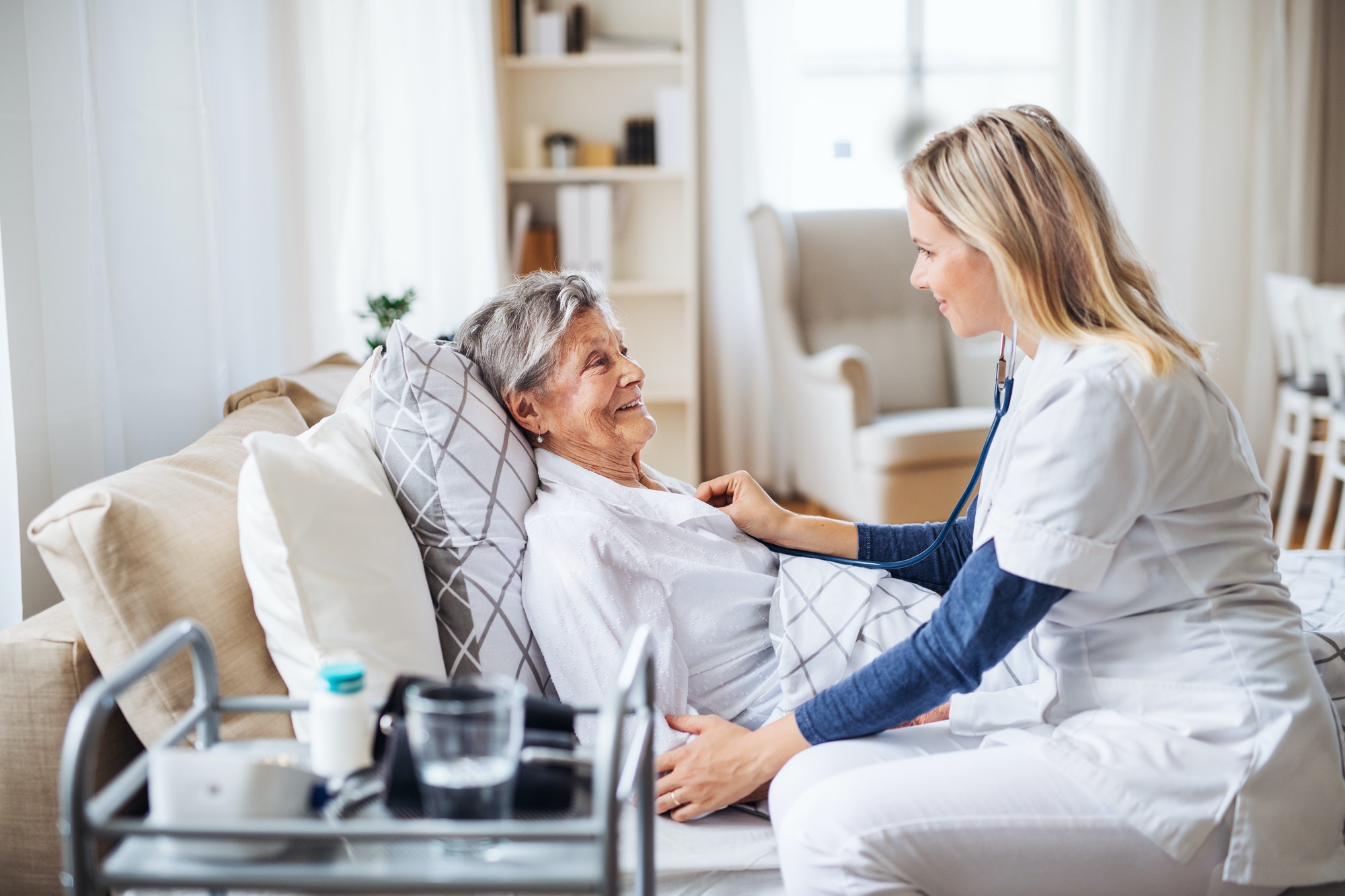 A health visitor examining a sick senior woman lying in bed at home with stethoscope.